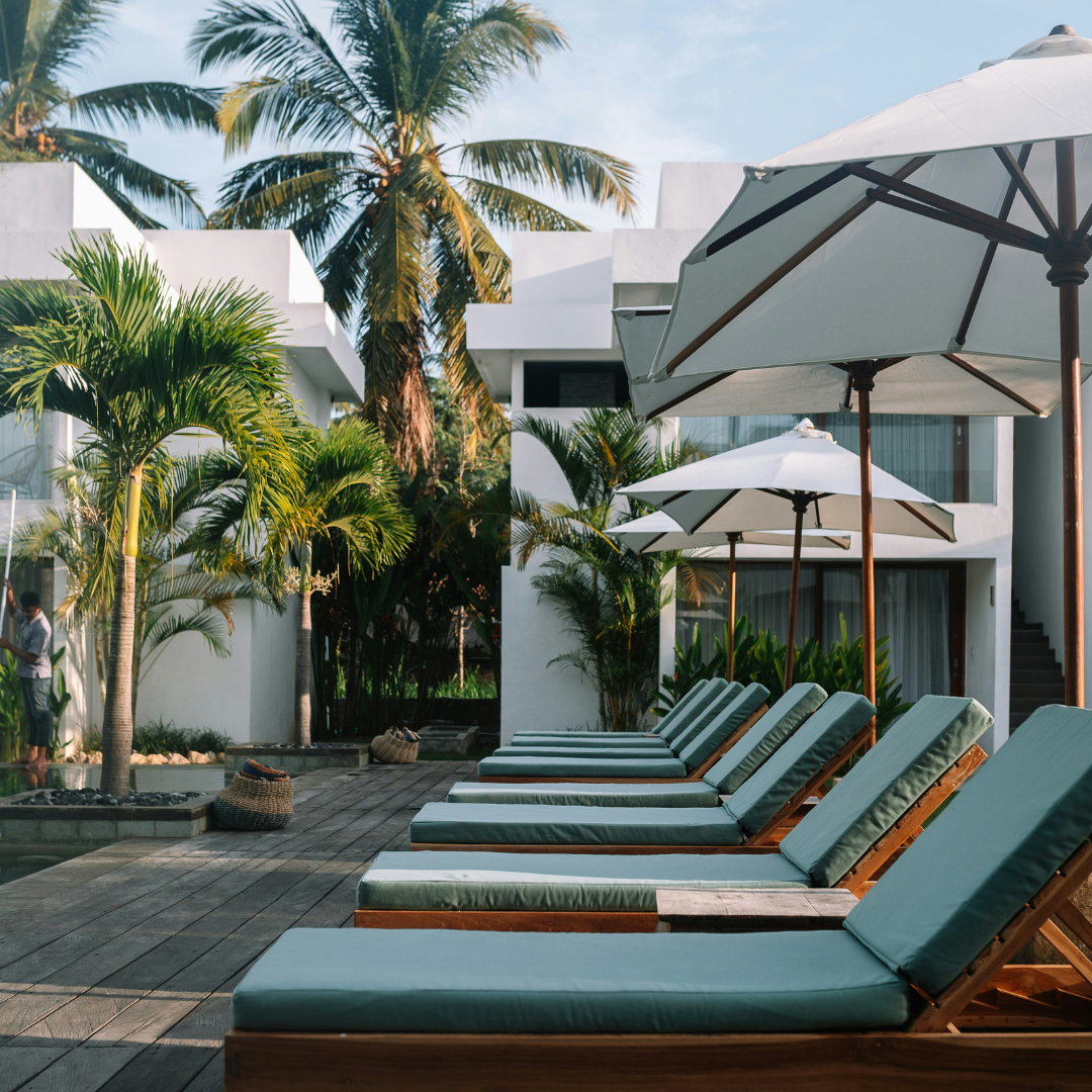 Row of green lounge chairs under white umbrellas with palm trees and a building in the background.